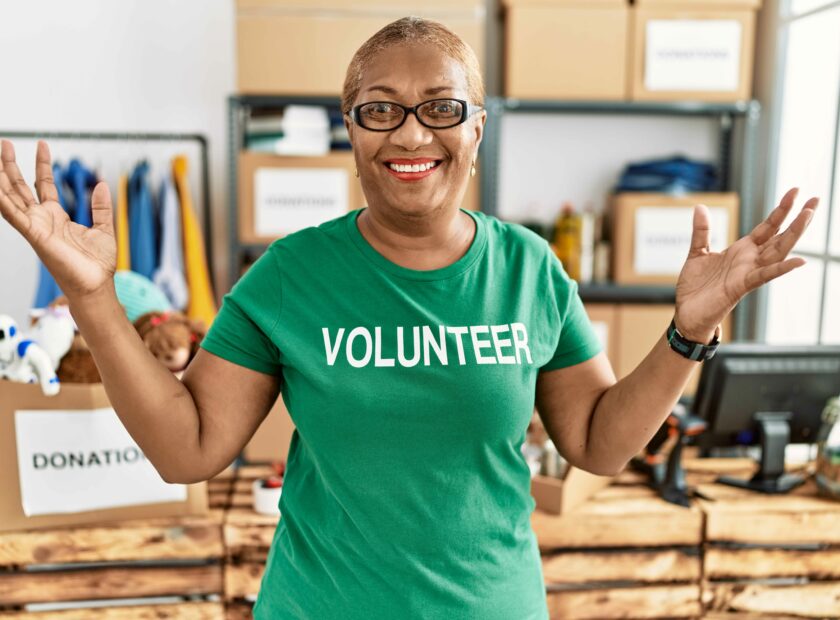 Mature hispanic woman wearing volunteer t shirt at donations stand celebrating victory with happy smile and winner expression with raised hands