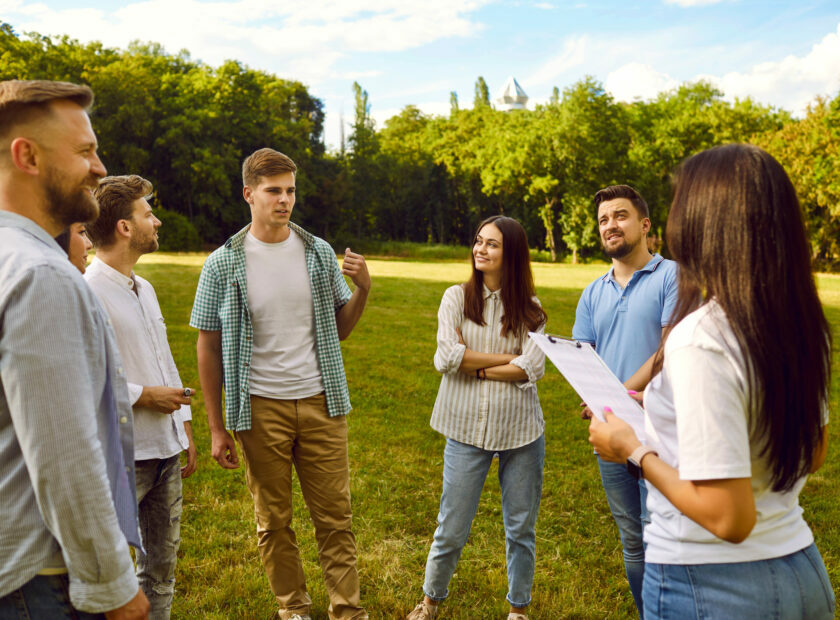Woman taking interview from a group of a young people standing in a circle in the park.