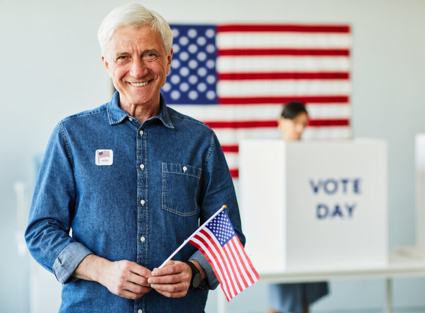 Waist up portrait of smiling senior man holding American flag in voting station on election day